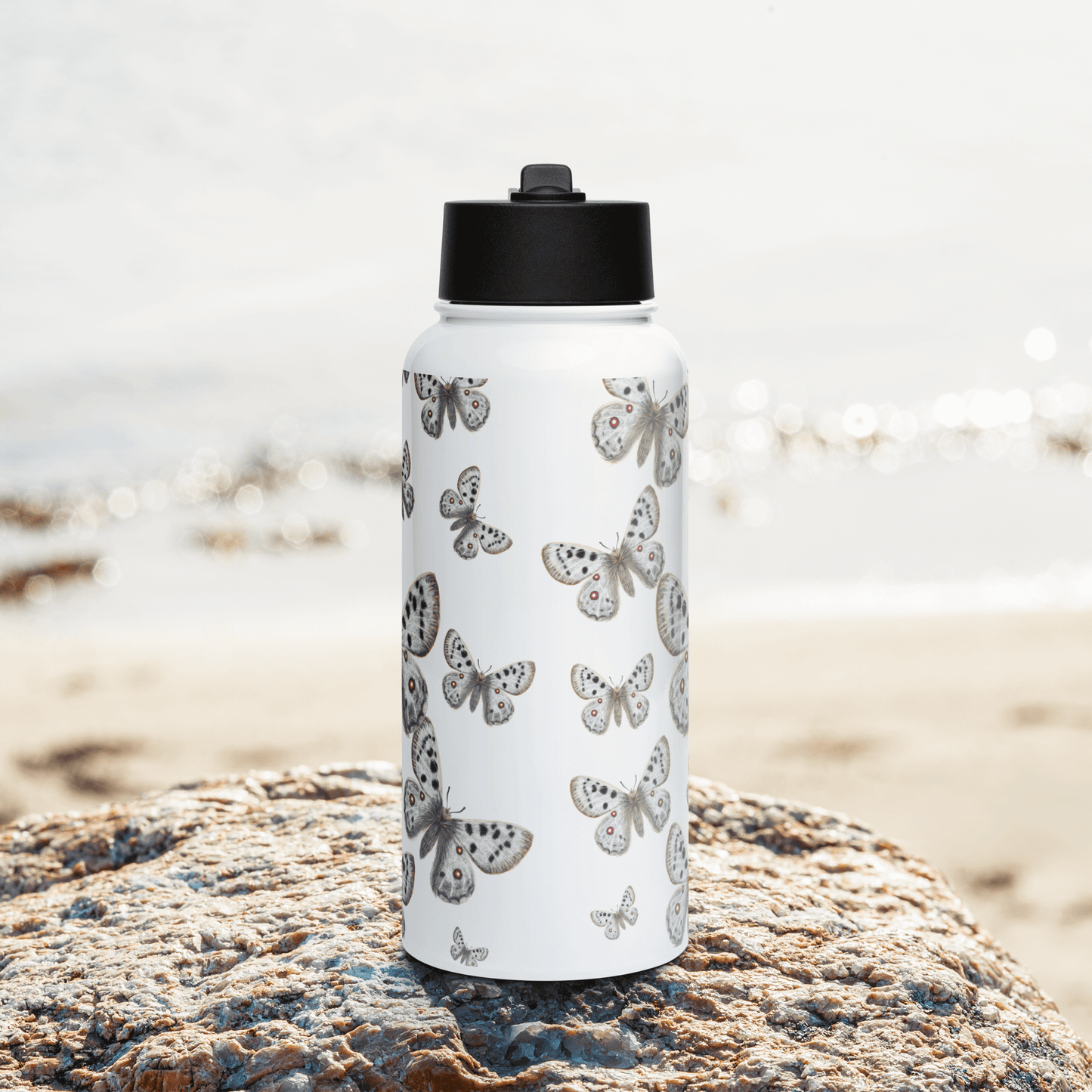 Elegant white stainless steel water bottle adorned with Apollo butterflies, placed on a rock by the beach.