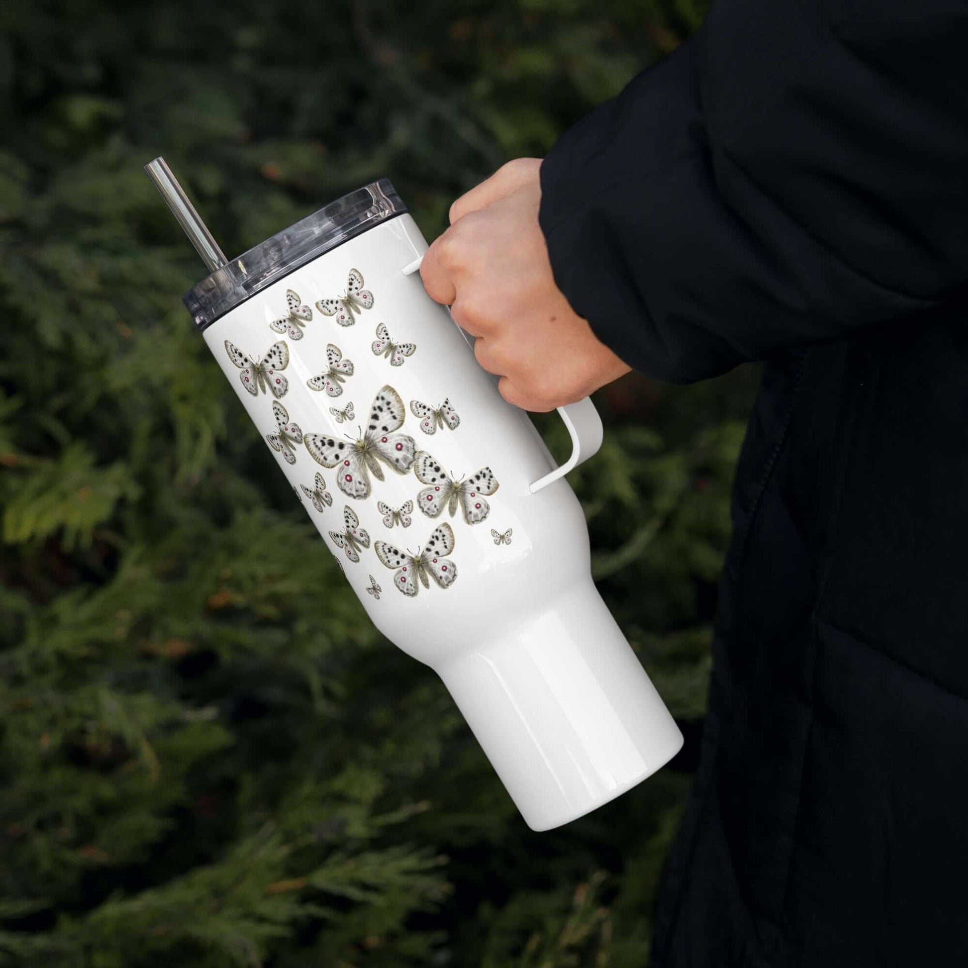 Apollo Butterfly Design Tumbler held by a person, featuring a sleek handle, lid, and straw amidst greenery.