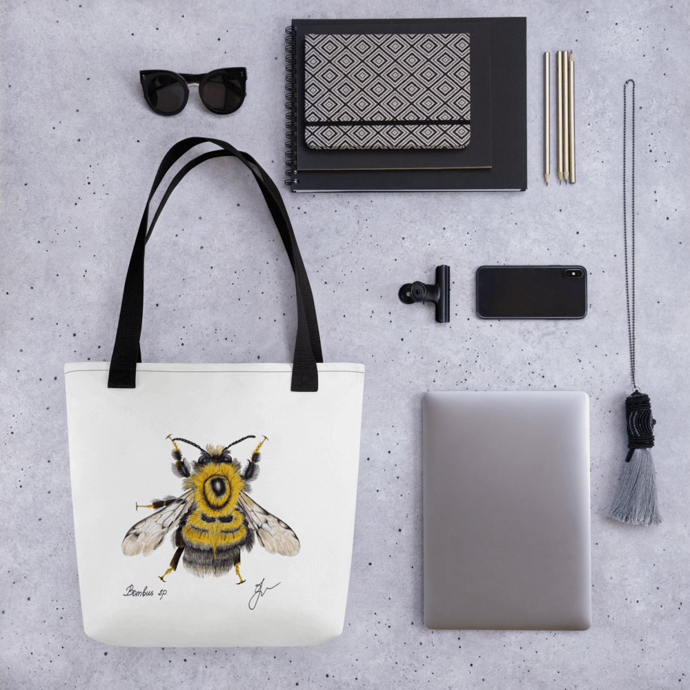 Bumblebee all-over print tote bag with black handles laying within multiple other products like glasses, mobile phones and notepads
