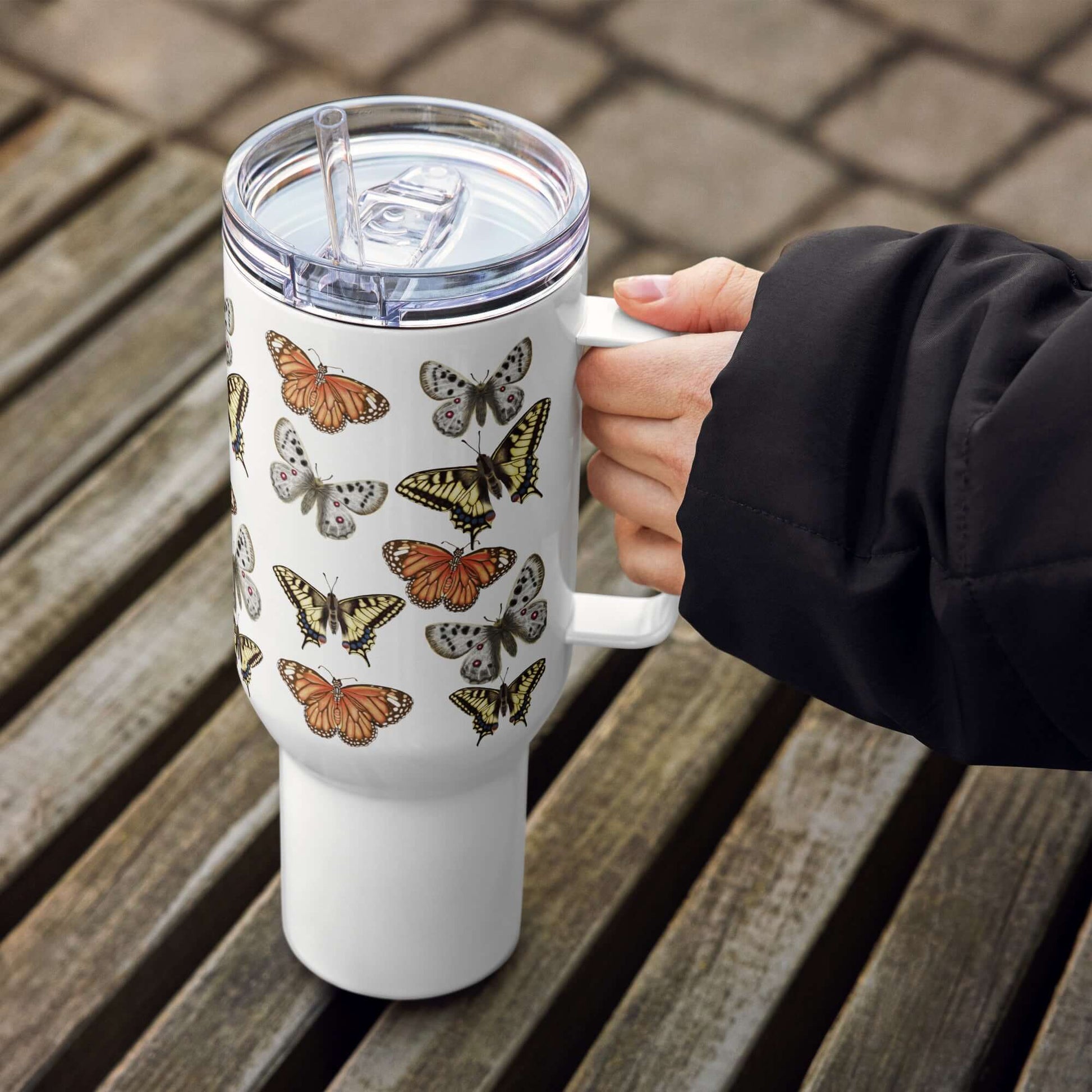 Butterflies design stainless steel tumbler with handle, held by a person on a wooden table, perfect for travel and drinks.