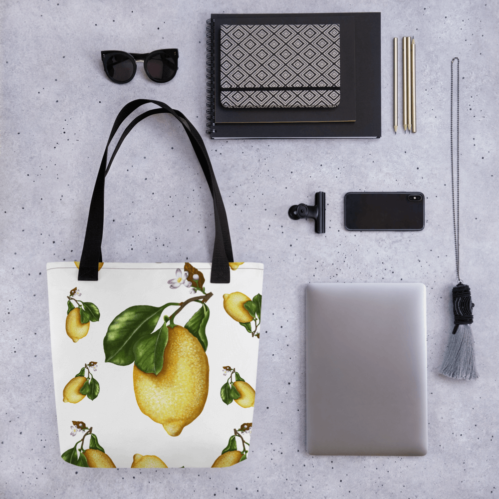 Lemon all-over print tote bag with black handles laying within multiple other products like glasses, mobile phones and notepads
