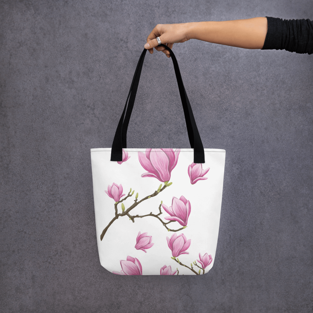 Magnolia all-over print tote bag with black handles held by a hand holding it straight
