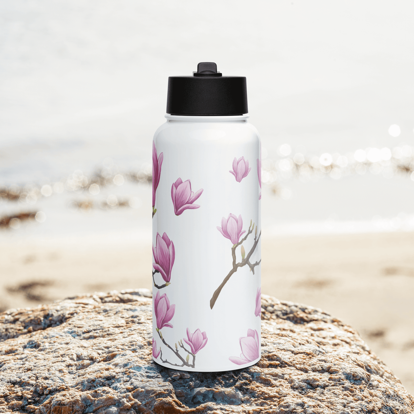 Elegant 32oz Magnolia water bottle displayed on a rock by the beach, featuring delicate pink blooms and a sophisticated design.