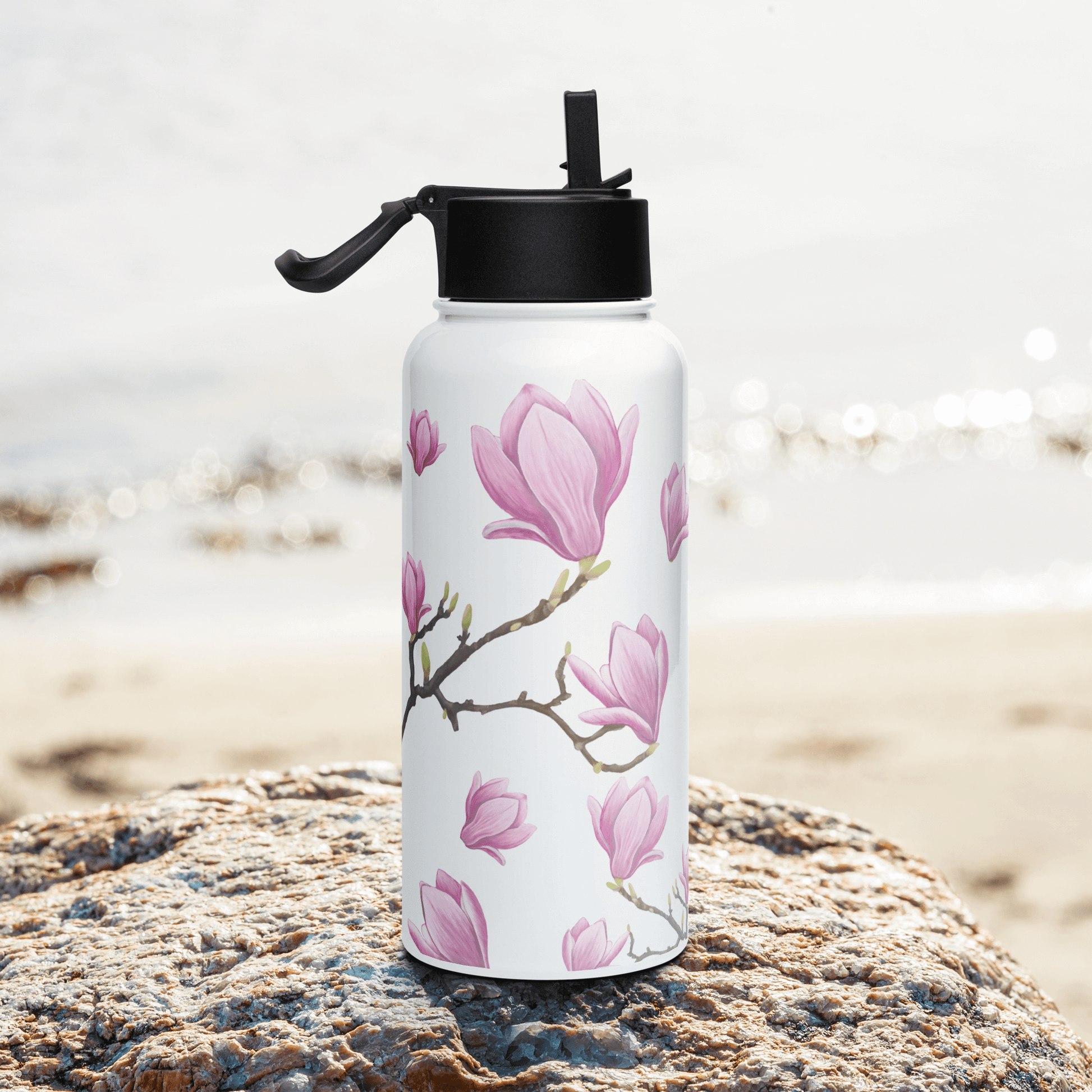 Elegant Magnolia water bottle on a rock by the beach, showcasing soft pink blooms and green leaves, perfect for gifting.