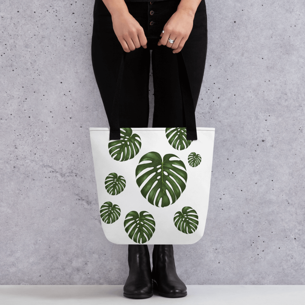 Monstera all-over print tote bag with black handles held by a hand holding it straight
