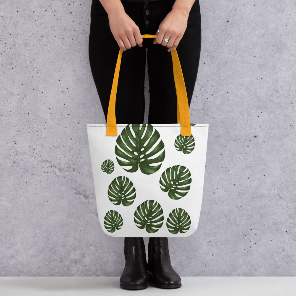 Monstera all-over print tote bag with yellow handles held by a hand holding it straight
