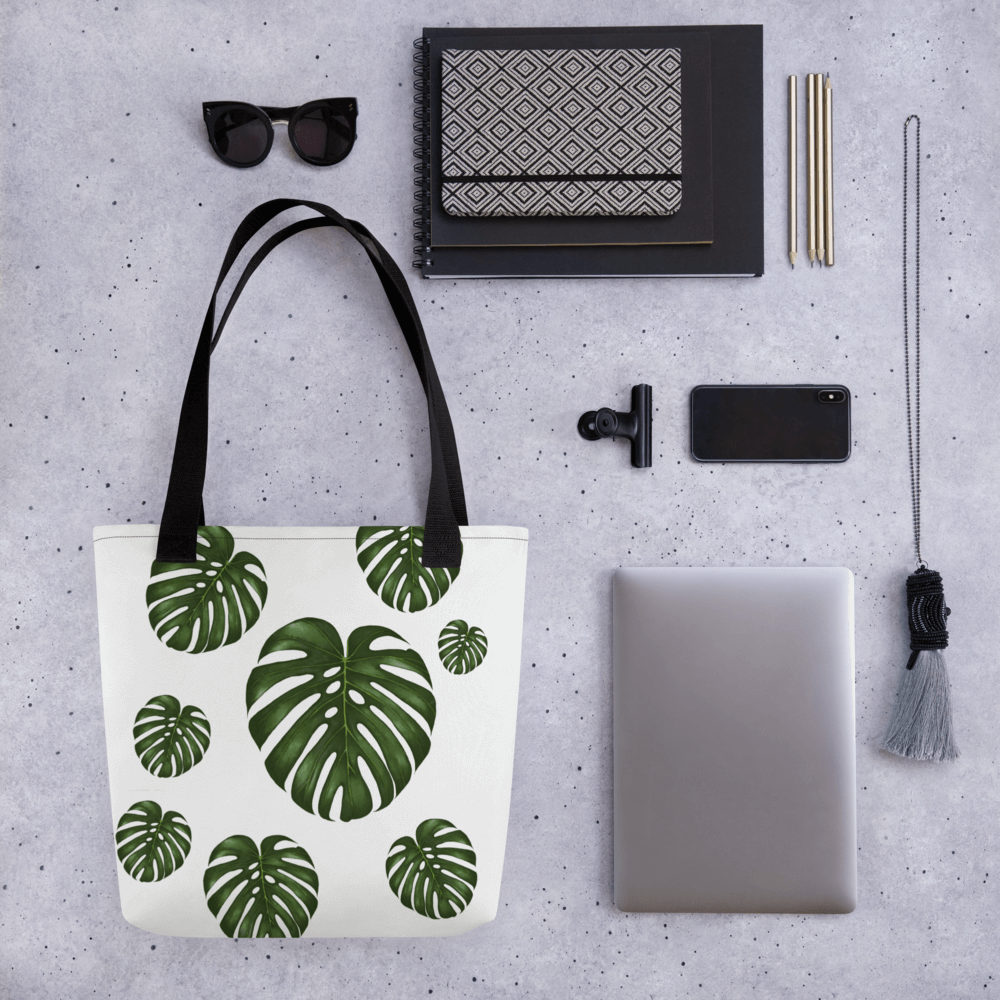 Monstera all-over print tote bag with black handles laying within multiple other products like glasses, mobile phones and notepads