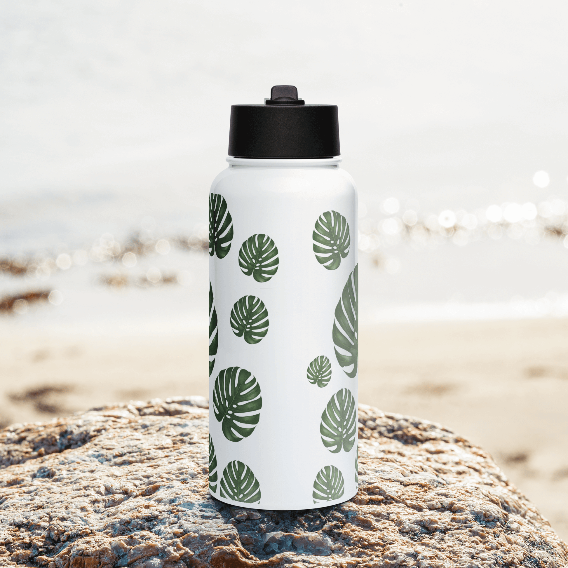 Tropical Monstera water bottle resting on a rock by the beach, showcasing botanical leaf design. Perfect gift for plant lovers.