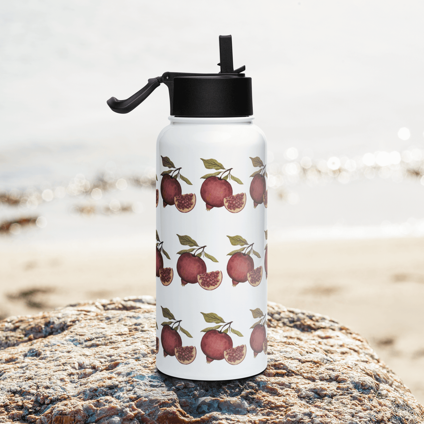 Vibrant pomegranate stainless steel water bottle on a beach rock, featuring a botanical design perfect for hydration.