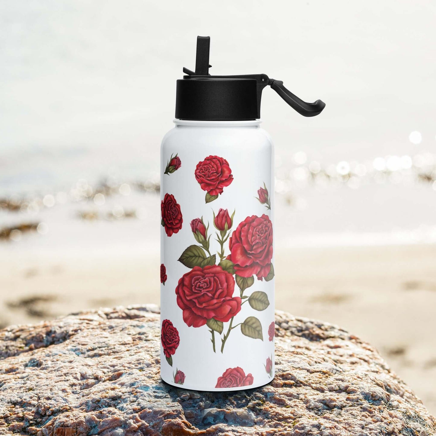 A 950ml rose water bottle rests on golden sand, its bright surface adorned with a romantic red rose design that contrasts beautifully with the blue ocean backdrop.