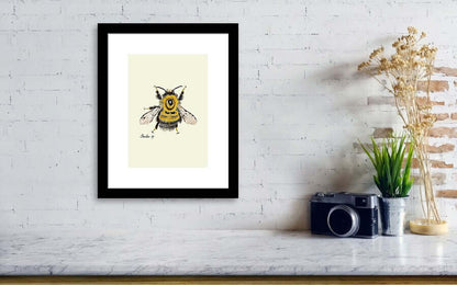 black framed bumblebeeart print hanging on a wall next to a vintage camera and a small vae green plant