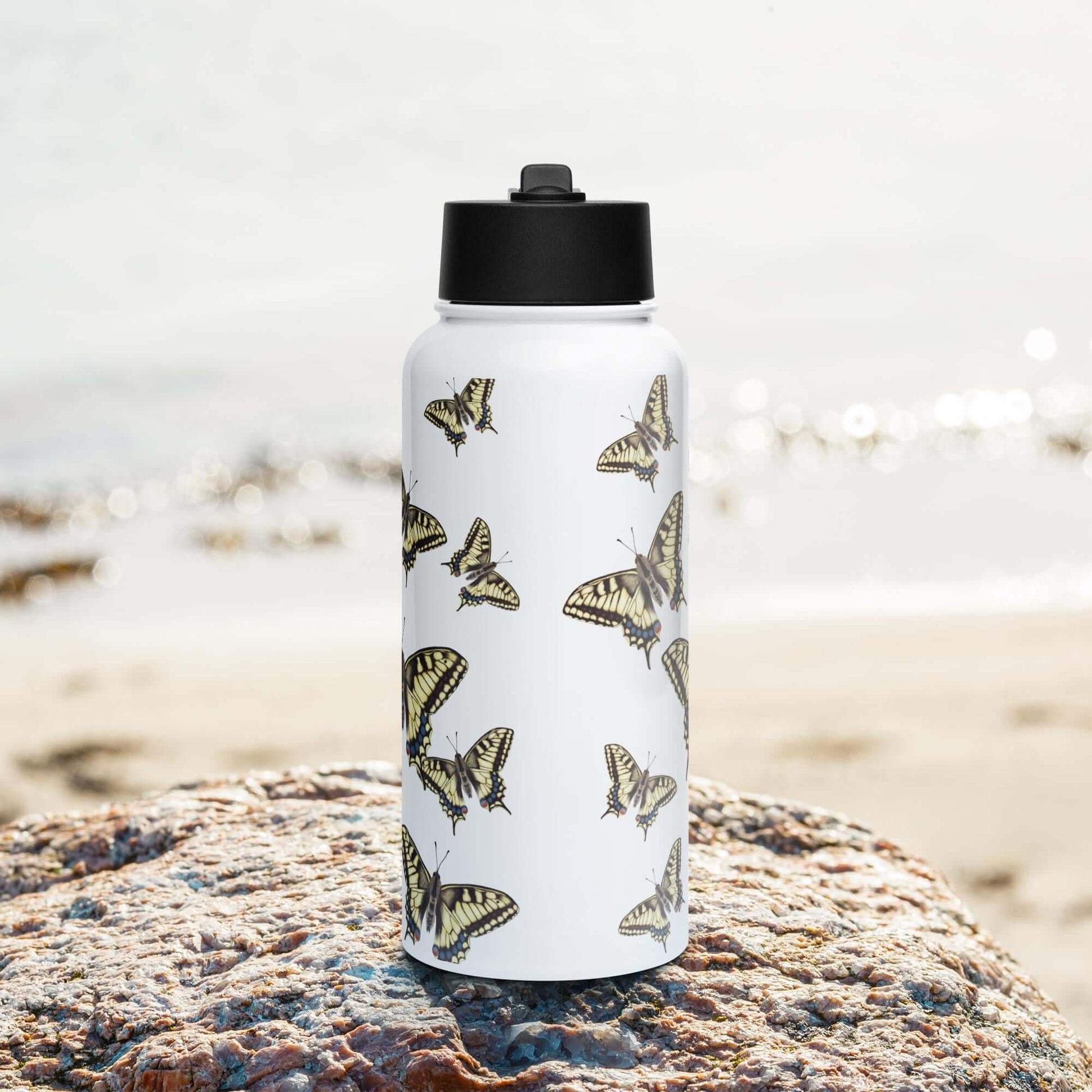 Front view on the beach: Elegant Machaon-inspired stainless steel water bottle with straw lid, perfect for a day at the beach.