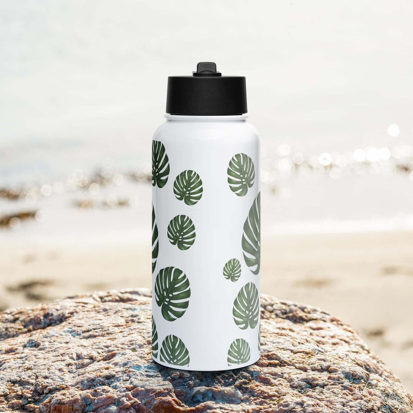 Front view on the beach: Tropical monstera-inspired stainless steel water bottle with straw lid, perfect for a day at the beach.