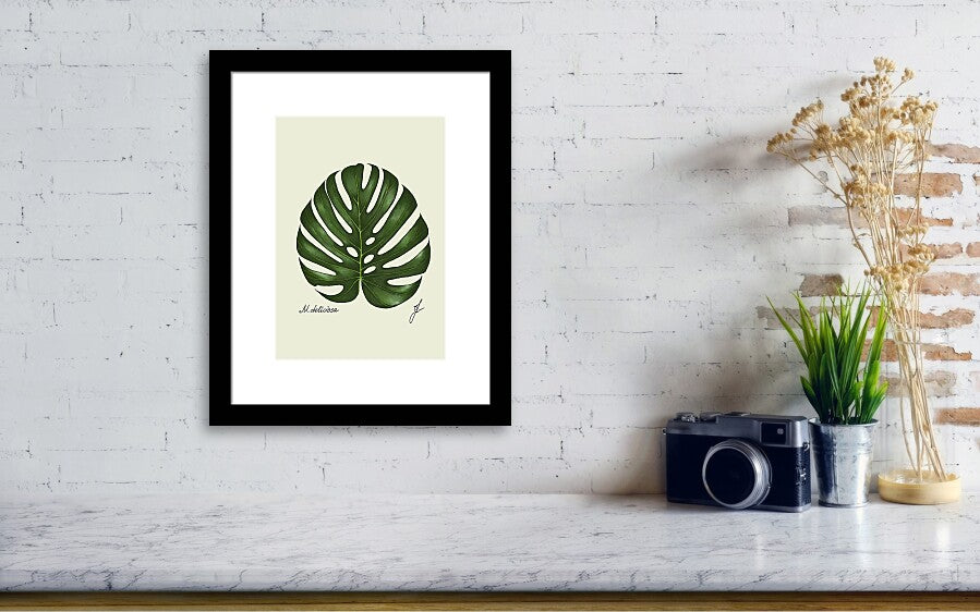 monstera black framed fine art print hanging on a wall next to a vintage camera and a small vae green plant